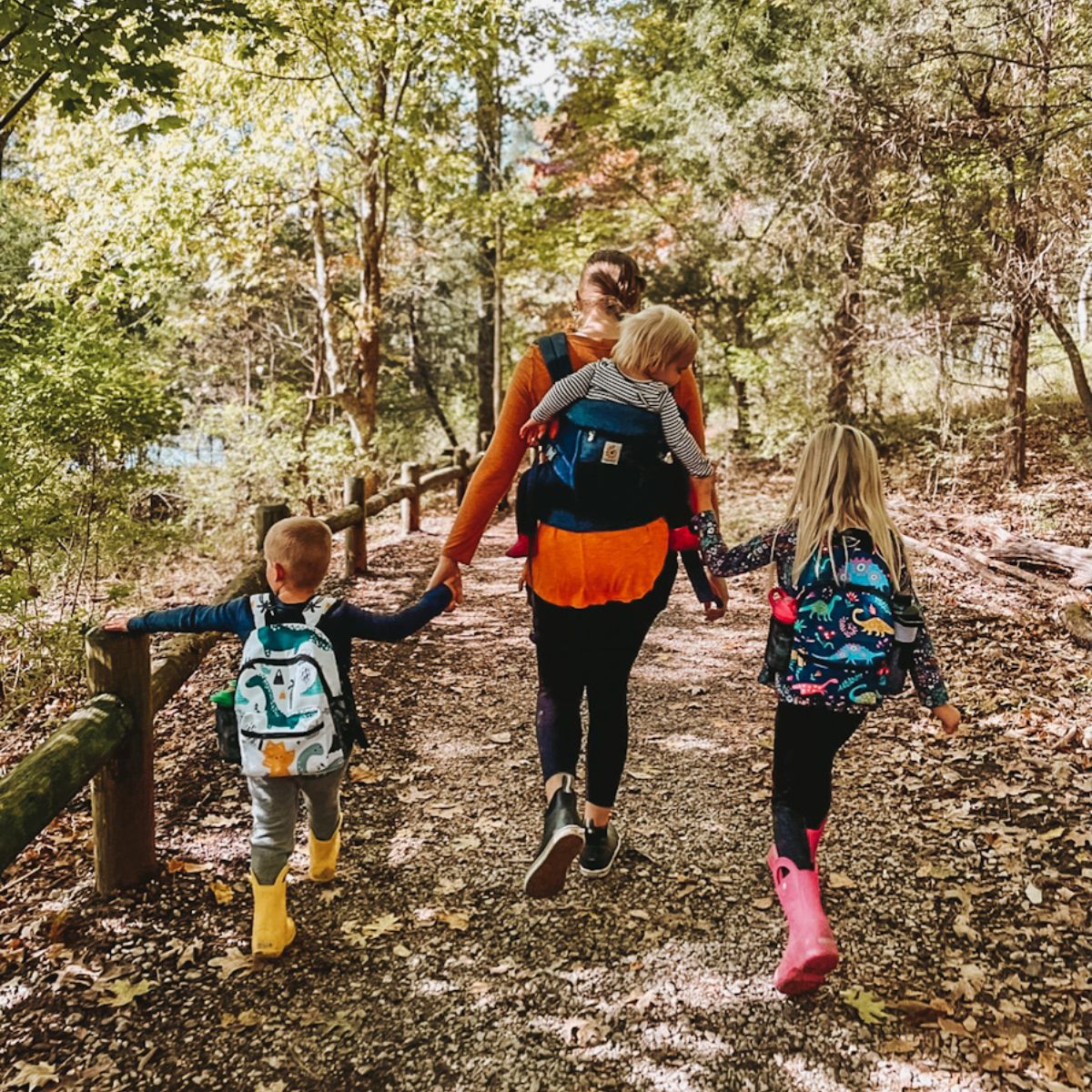 mom wearing toddler on back and holding hands with two other kids while hiking