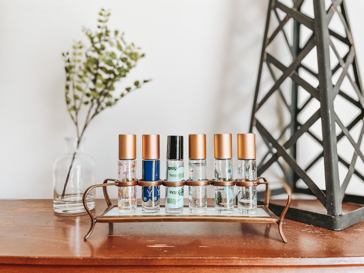 essential oil roller bottles on top of a wooden surface with a mini glass vase with greenery and a metal tower next to it
