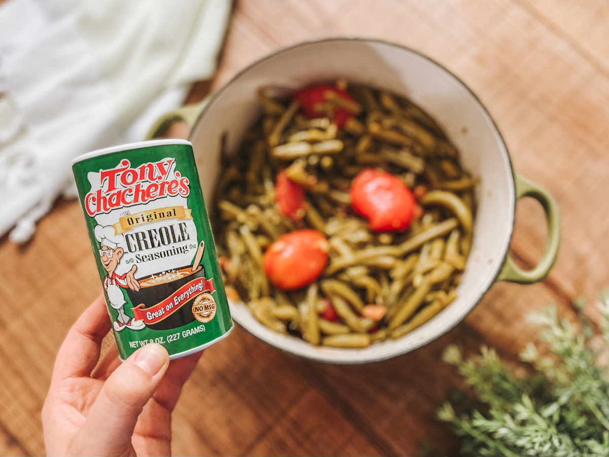 Tony Chachere's creole seasoning held up in front of a pot of green beans with tomatoes and bacon