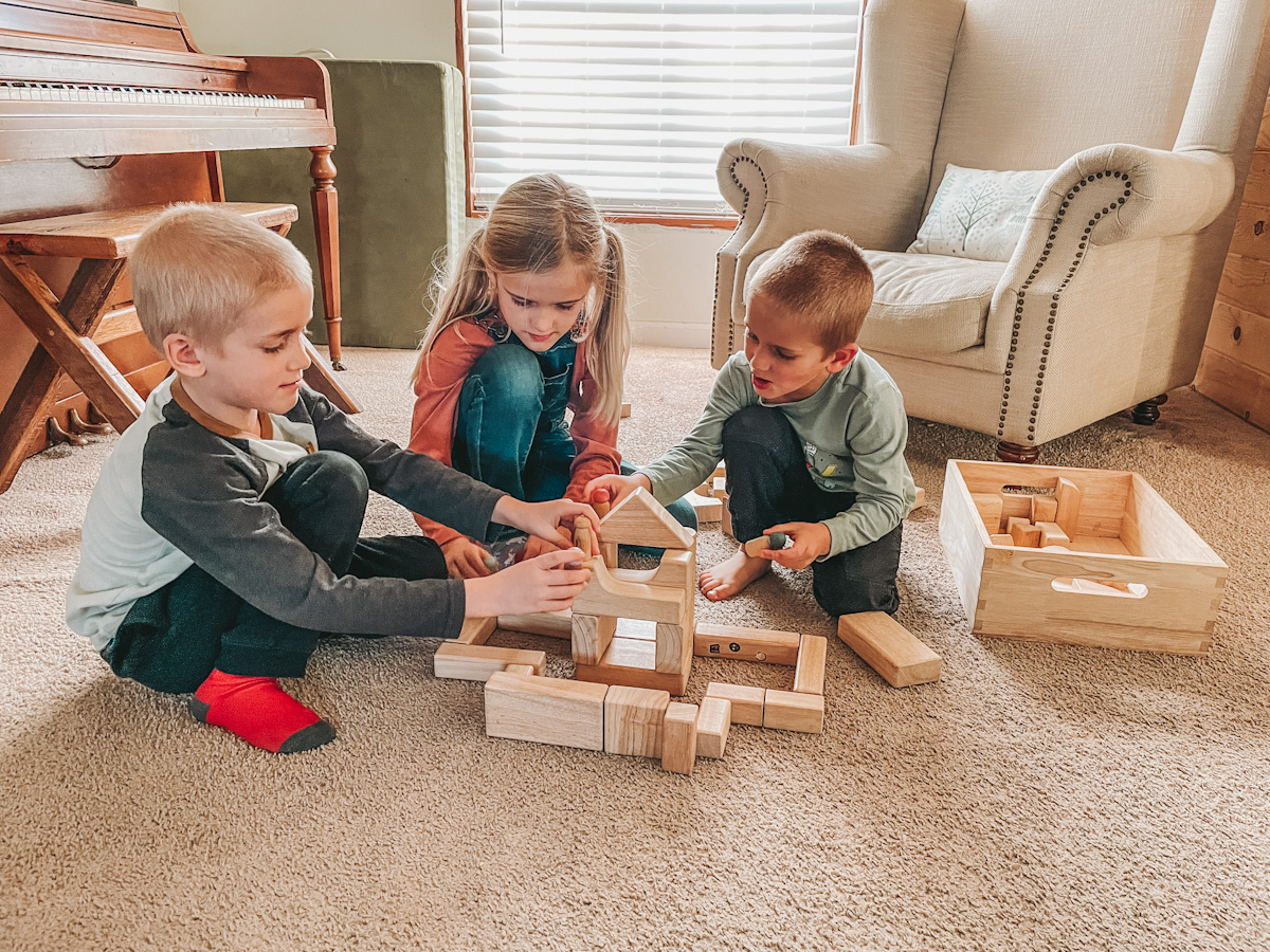 three kids sitting on the floor playing with open ended wood blocks and peg dolls