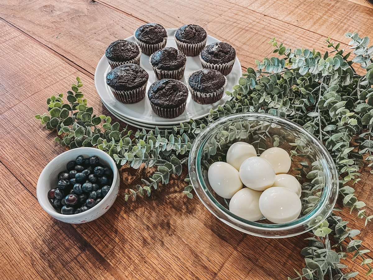 chocolate muffins, boiled eggs and blueberries in bowls laid out on a wood countertop