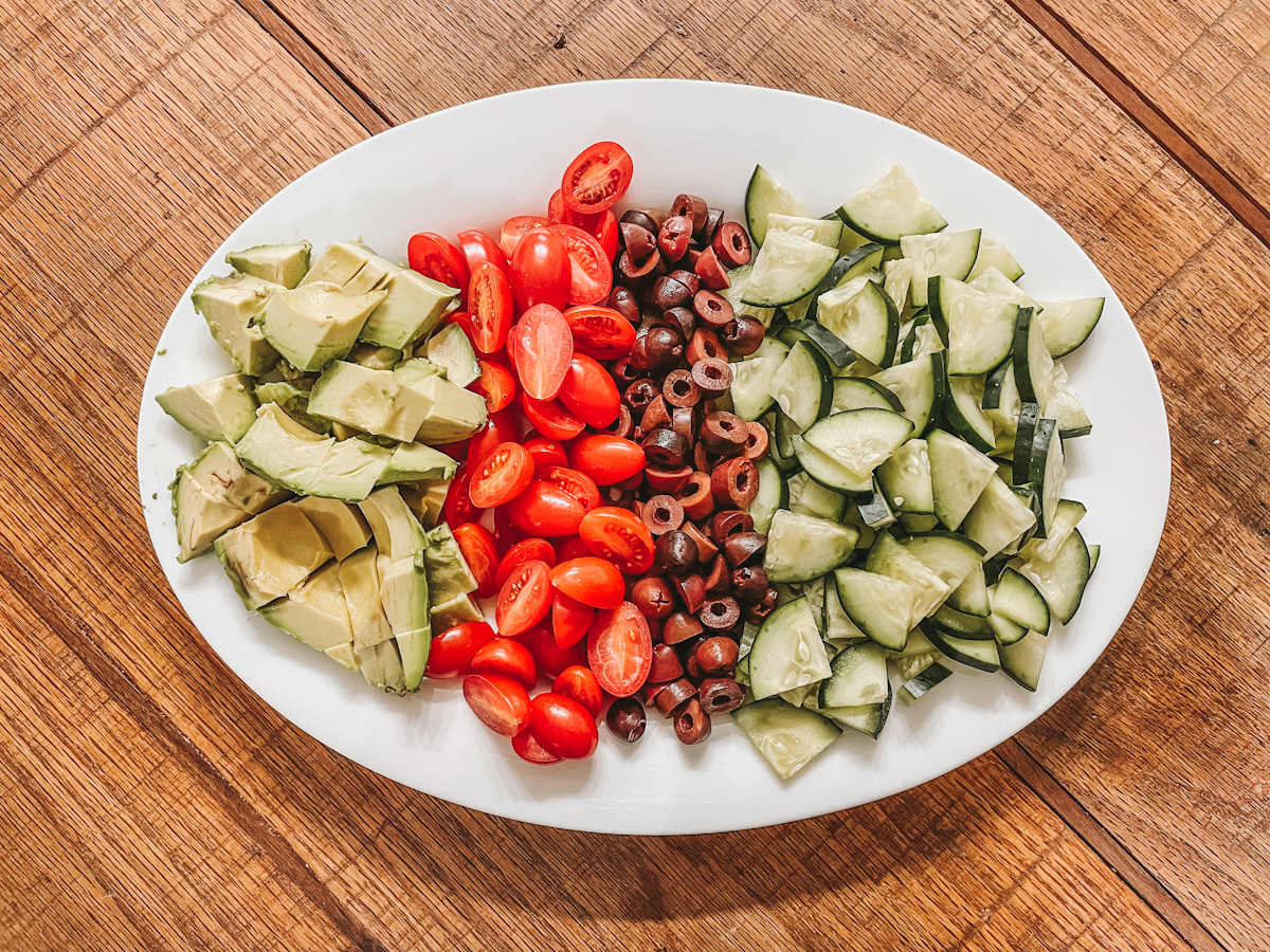 a white platter filled with avocado, cherry tomatoes, kalamata olives, and cucumber