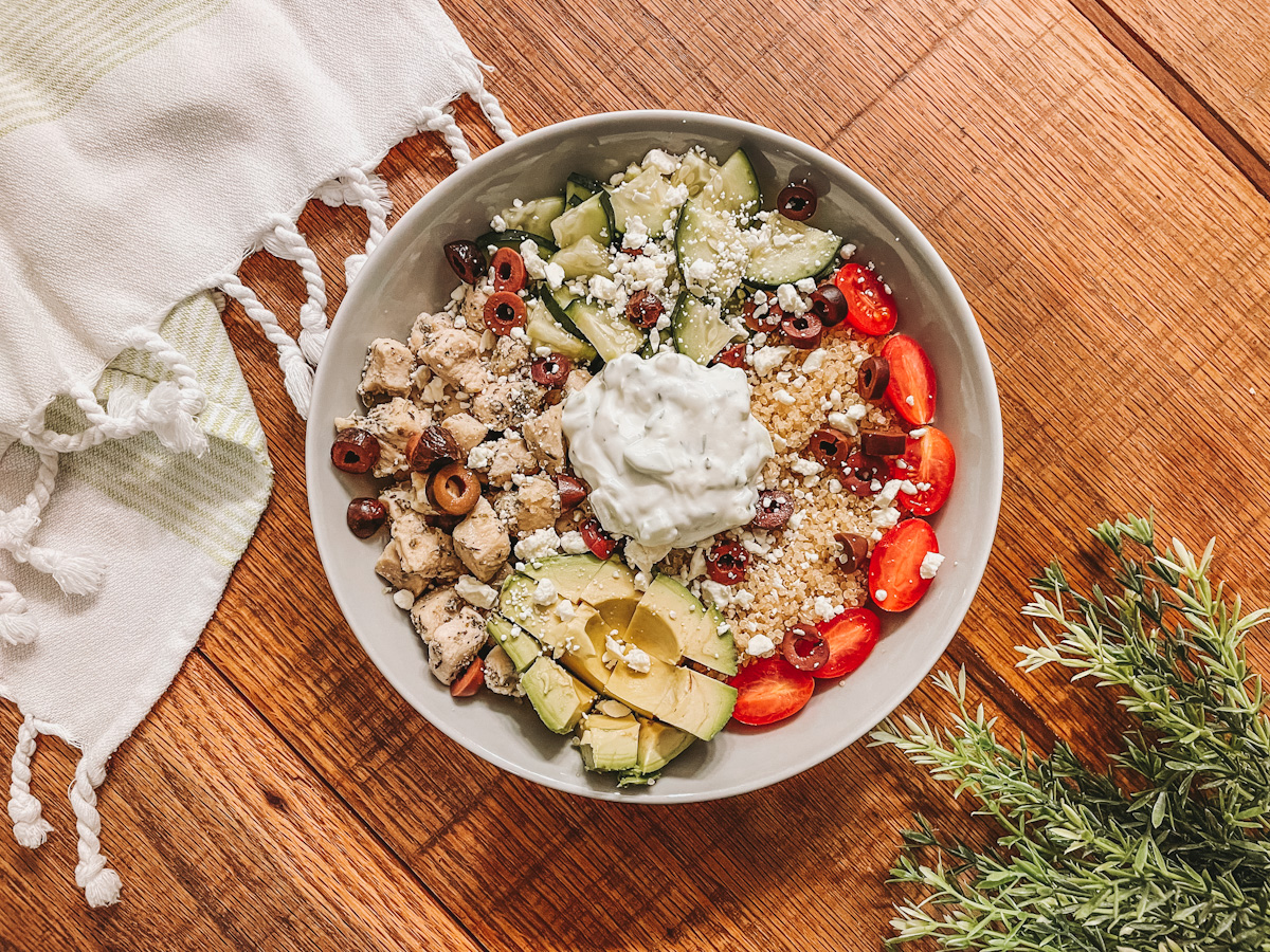 Greek chicken with quinoa, cucumber, avocado, kalamata olives, feta, topped with Tzatziki served in a white bowl