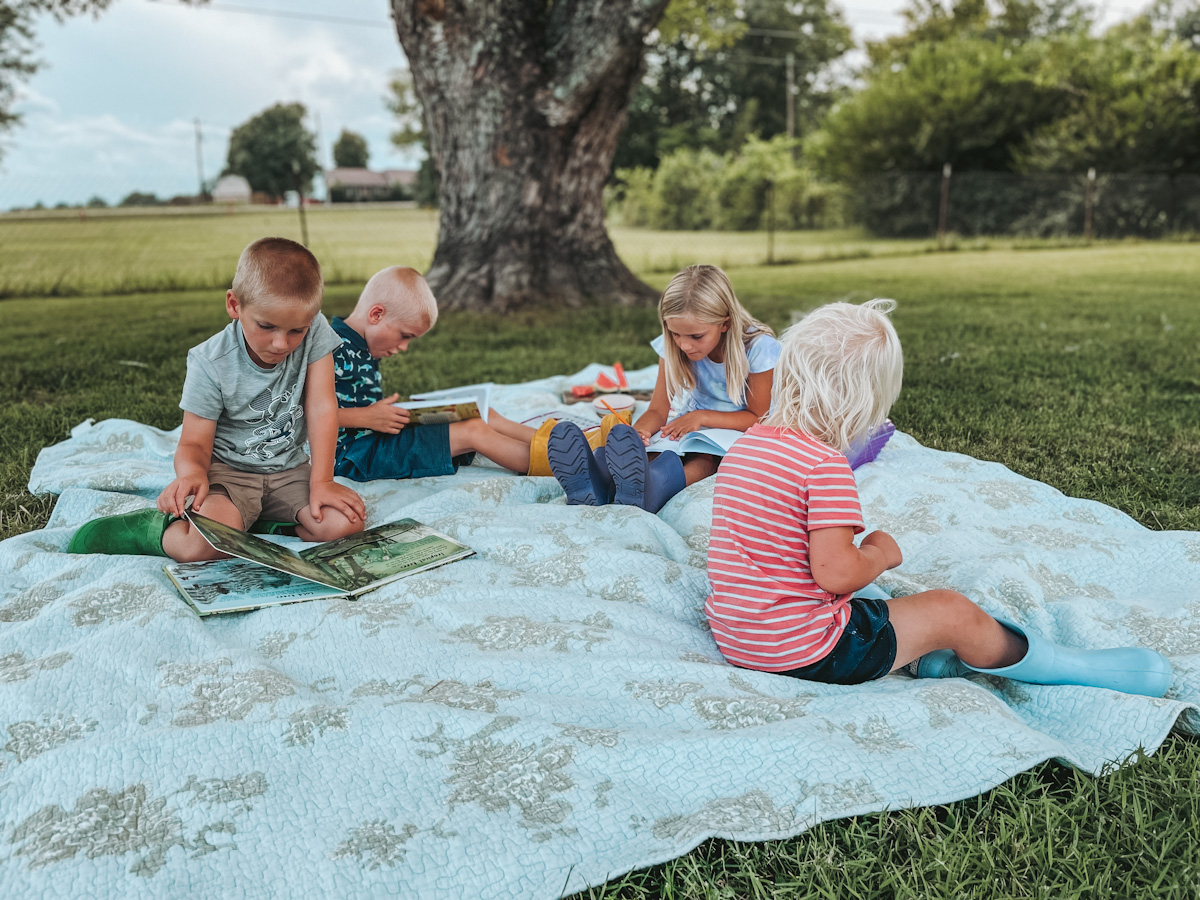 four homeschool kids sitting outside on a picnic blanket, looking at books