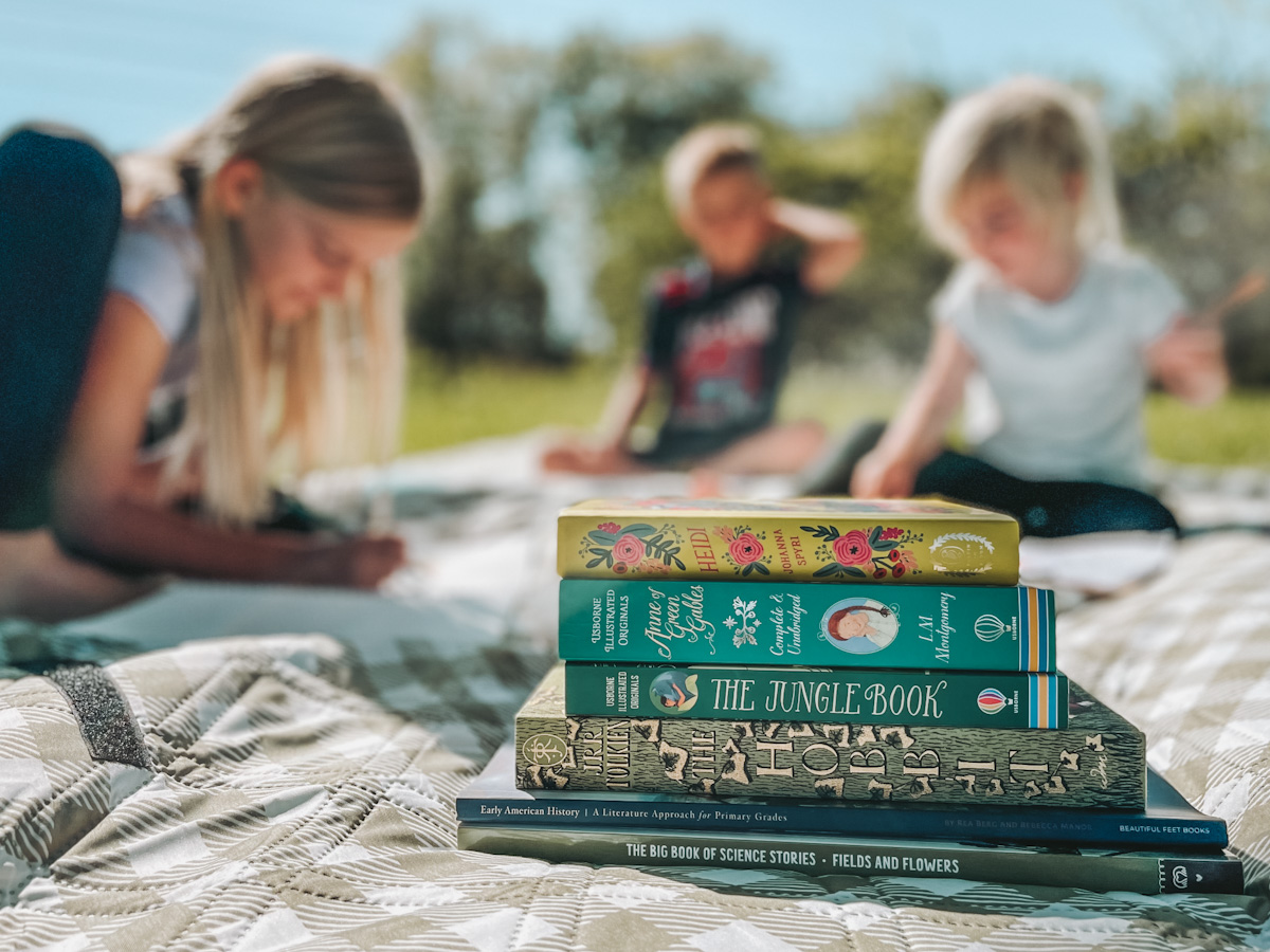 a summer book stack in front of 3 kids drawing and coloring on a picnic blanket for their homeschool schedule