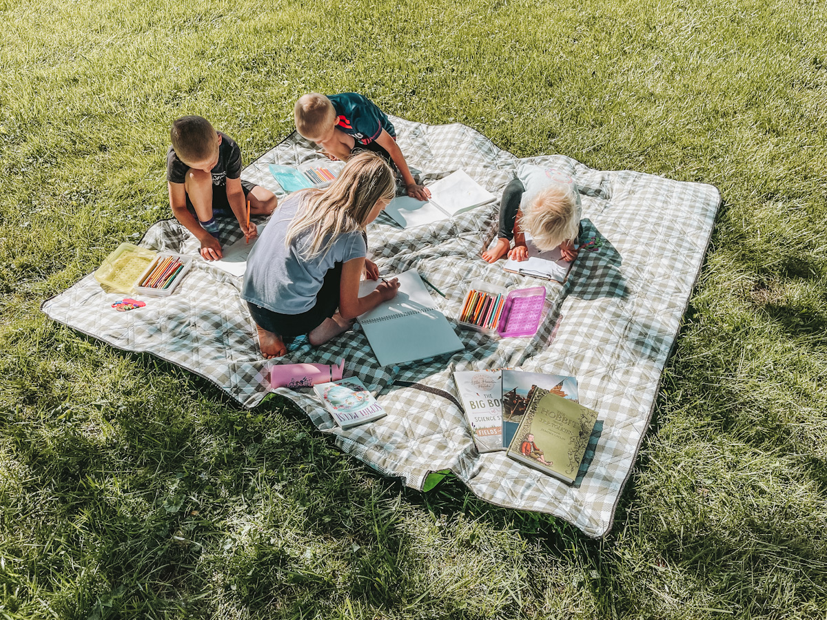 a green and white checkered picnic blanket spread out on the grass, with 4 kids sitting on it drawing and coloring for their summer homeschool schedule