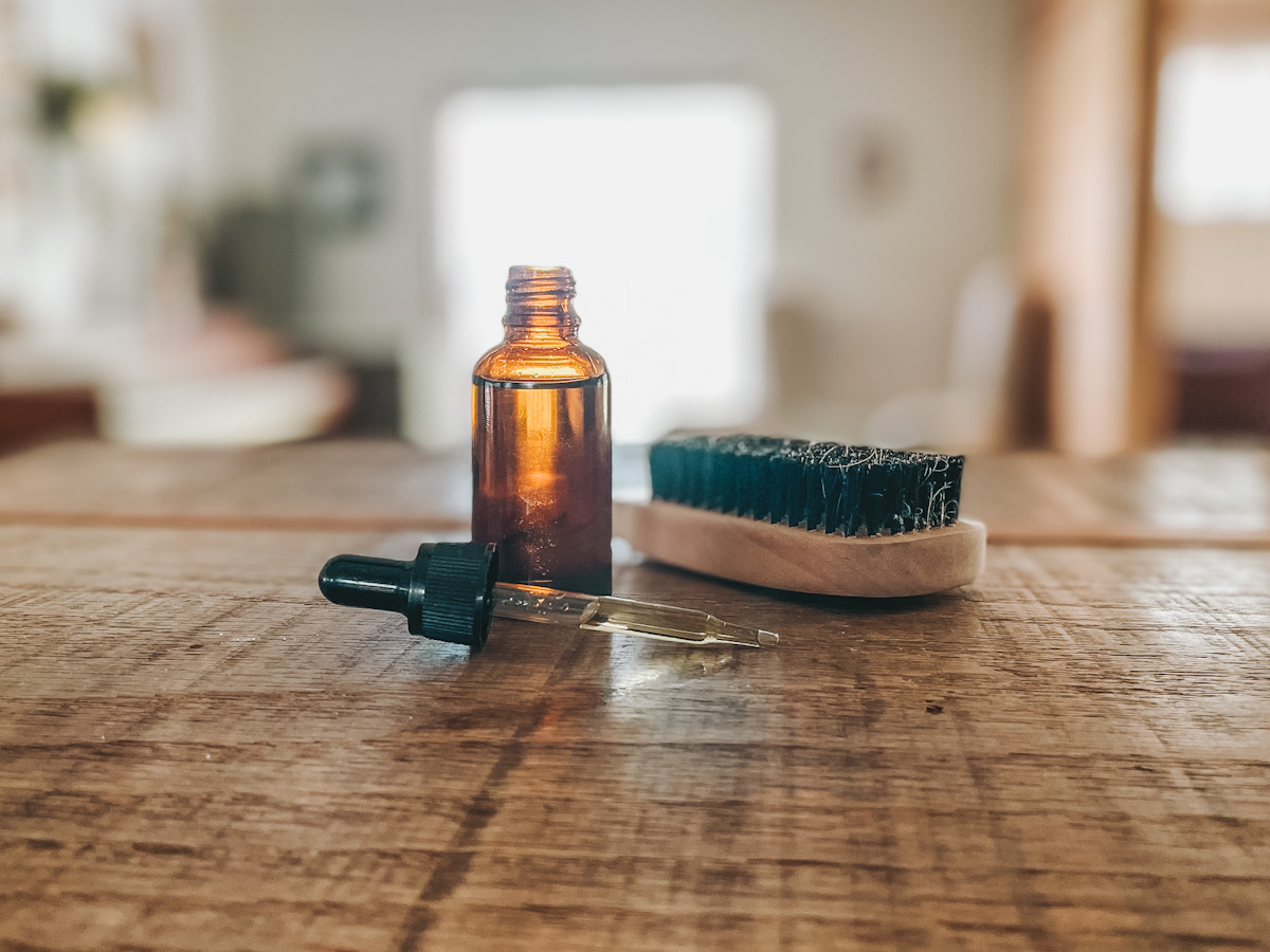 amber glass bottle set on a wood countertop with a beard brush beside it and a dropper in front filled with DIY beard oil