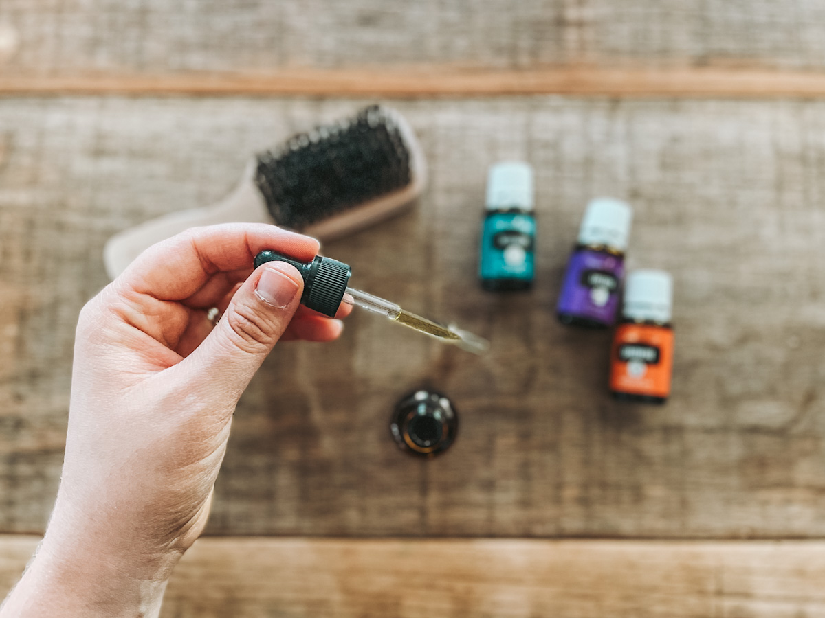 hand holding a dropper filled with DIY beard oil above a wood countertop with essential oil bottles and a beard brush