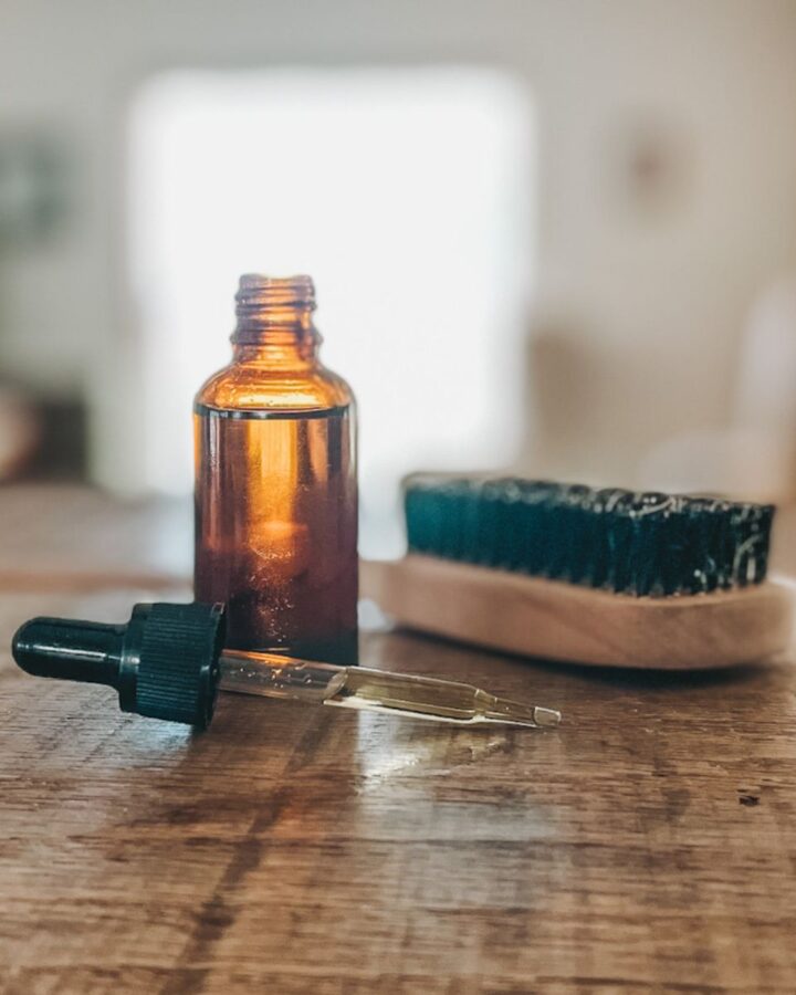 amber glass bottle on a wood countertop with a beard brush beside it and a dropper in front filled with DIY beard oil